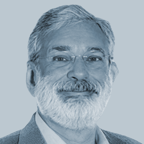 Portrait of a smiling older man with gray hair and beard, wearing glasses and a blazer.