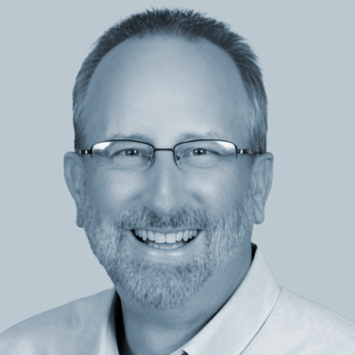 Portrait of a smiling man with short gray hair and glasses, wearing a light shirt against a pale blue background.