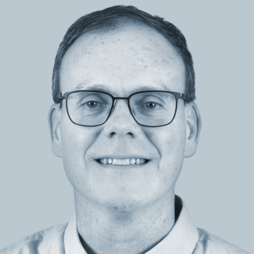 Smiling man with short hair and rectangular glasses facing the camera against a light blue background.